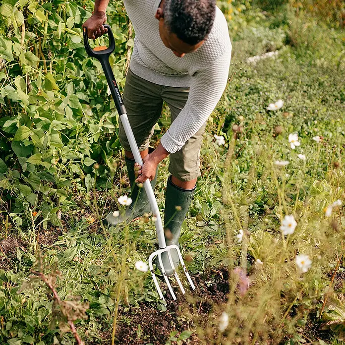Fiskars Xact Spatengabel - Arbeitsbreite: 18,5 cm, Anzahl Zinken: 4, Teleskopstiel - Der Fiskars Grabegabel: Ein Mann verwendet sie in seinem Garten zum Auflockern des Bodens, umgeben von üppigem Grün und Blumen.
