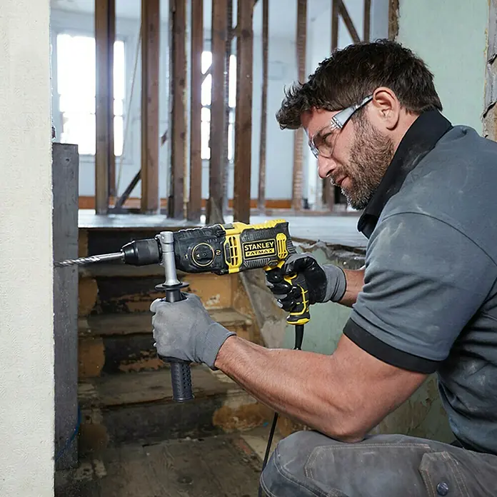 Stanley FatMax Taladro percutor FMEH1100K con maletín - 1.100 W, Portabrocas de sujeción rápida - Un artesano usa un martillo rotativo Stanley Fatmax amarillo y negro para perforar una pared, mientras usa gafas de seguridad y guantes.