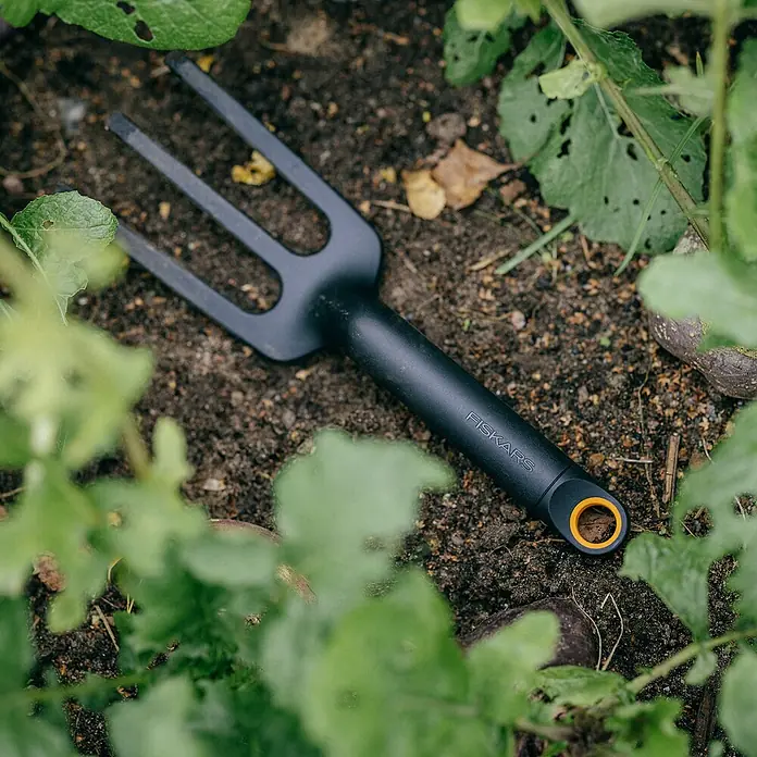 Fiskars Solid Hand-Kleinrechen - 27,7 cm - Nahaufnahme einer schwarzen Fiskars Gartenkralle auf Erde, umgeben von grünen Pflanzen. Perfekt für Gartenarbeiten und zum Auflockern des Bodens.