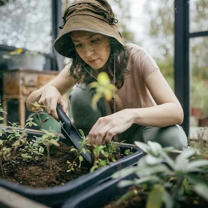 Fiskars Solid Pflanzkelle - 28,2 cm - Eine Frau mit Hut, die in einem Garten mit einer Gartenschaufel in der Erde arbeitet und junge Pflanzen setzt.