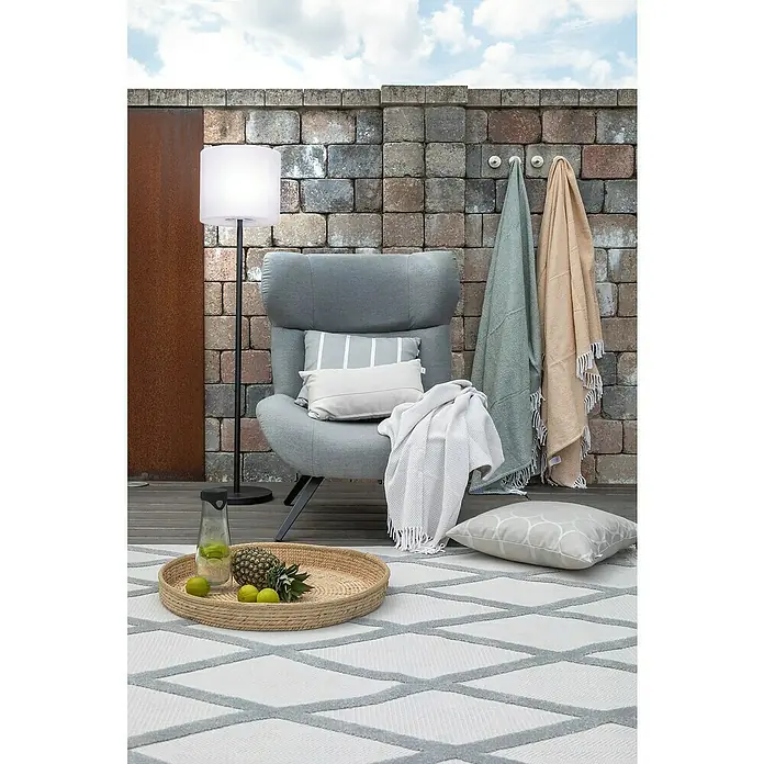 SCHÖNER WOHNEN-Kollektion Akku-Stehleuchte MINA - 34 x 34 x 150 cm, Sandschwarz, RGB, 2 W, IP44 - Gartenszene mit grauem Sessel, Stehlampe und Outdoor-Teppich mit geometrischem Muster, dekoriert mit Kissen und Decken für eine gemütliche Atmosphäre.