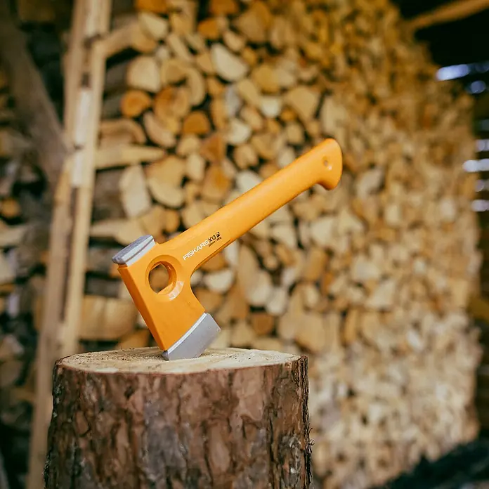 Fiskars X-series Universalaxt X13 - Länge: 33,2 cm - Nahaufnahme einer orangefarbenen Fiskars X13 Spaltaxt, die in einen Holzscheit gesteckt ist, mit einem Stapel Holz im Hintergrund.