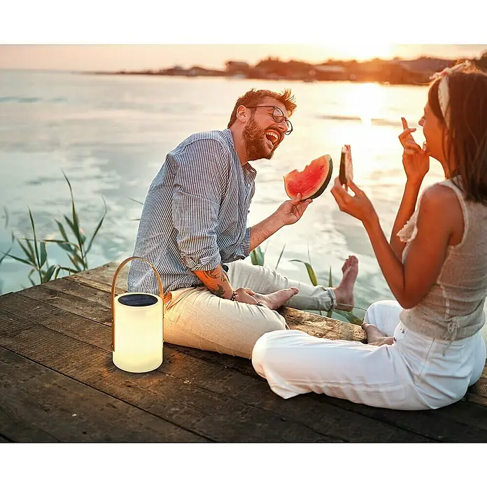 FHL easy Akku-Tischleuchte BARI - 0,5 W, L x B x H: 16 x 16 x 34 cm, Weiß, RGB - Idyllische Szene mit einem Paar am Seeufer, beleuchtet von einer sanften Laterne, die eine warme Atmosphäre schafft.\n