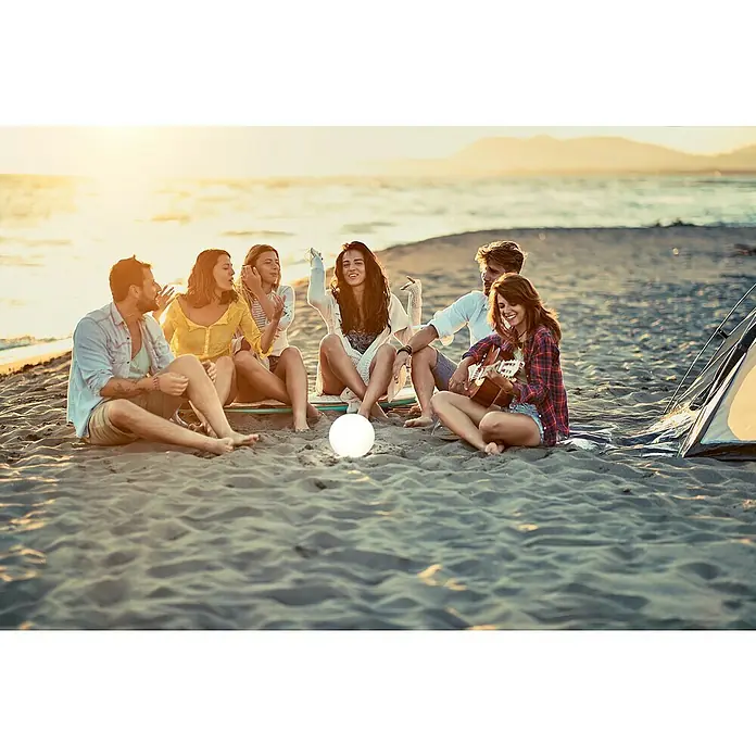 FHL easy Akku-Tischleuchte BUSSO - 0,1 W, L x B x H: 18 x 18 x 16 cm, Weiß, RGB - Eine Gruppe Freunde sitzt am Strand mit einer Leuchtkugel, eine Frau spielt Gitarre am Lagerfeuer mit Zelt im Hintergrund.