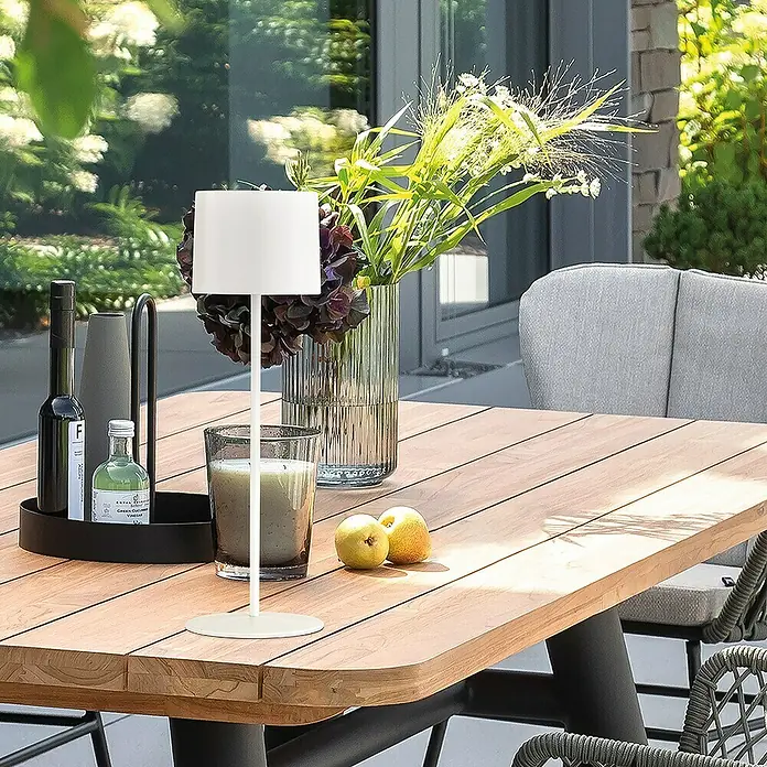 SCHÖNER WOHNEN-Kollektion Akku-Tischleuchte SUN - 2 W, L x B x H: 12 x 12 x 38 cm, Sandweiß, RGB - Nahaufnahme eines Tisches mit einer modernen weißen Lampe, einer Vase mit Blumen und einer Flasche Öl, ideal für den Außenbereich.