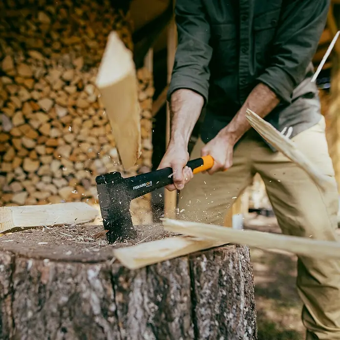 Fiskars X-series Universalaxt X24 M - Länge: 60 cm - Ein Mann spaltet Holz mit einer Fiskars Axt. Die Axt mit dem orangefarbenen Griff ist im Einsatz, Holzsplitter fliegen durch die Luft.