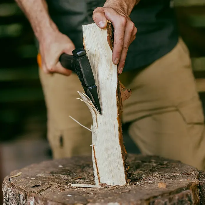 Fiskars X-series Universalaxt X14 - Länge: 35,5 cm - Ein Mann spaltet Holz mit einer Axt auf einem Baumstumpf. Das Holzstück ist fast vollständig gespalten und zeigt die Holzfasern.