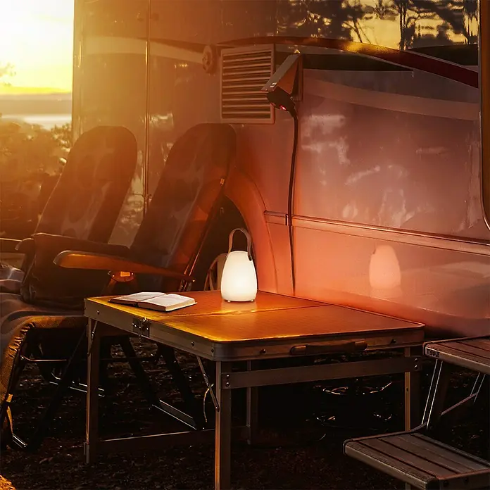 FHL easy Akku-Tischleuchte RUBY - 0,5 W, L x B x H: 12 x 12 x 16 cm, Weiß, RGBW, 1 Stk. - Camping-Szene mit Tisch und leuchtender Laterne im Vordergrund, umgeben von Stühlen und Wohnmobil, für gemütliche Outdoor-Beleuchtung.