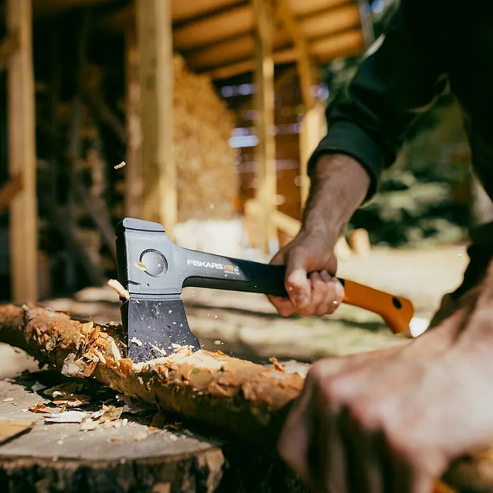 Fiskars X-series Universalaxt X18 - Länge: 44,5 cm - Eine Axt der Marke Fiskars spaltet einen Holzstamm in einer rustikalen Umgebung, Holzspäne fliegen.