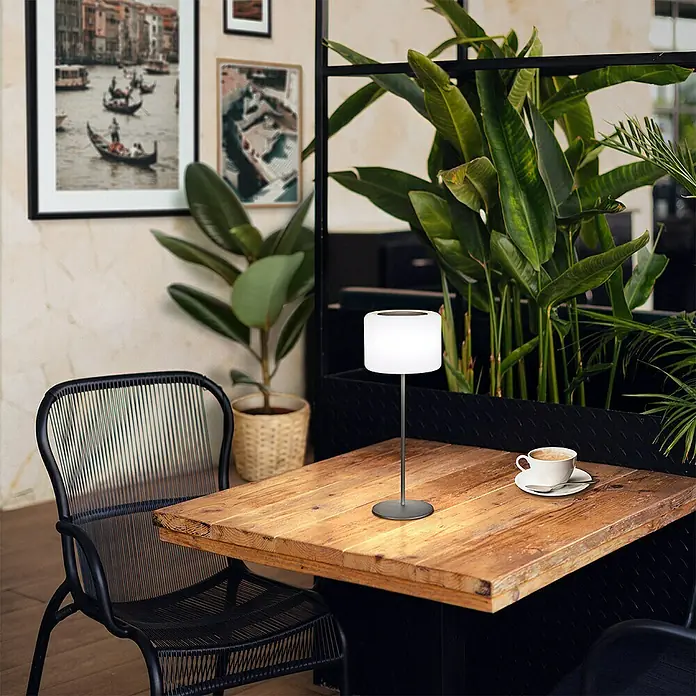 FHL easy Akku-Tischleuchte PINO - 0,5 W, L x B x H: 15 x 15 x 39,5 cm, Sandschwarz, RGB - Eine moderne Tischleuchte mit einem zylindrischen Lampenschirm steht auf einem Holztisch. Die Lampe ist ideal für gemütliche Abende.