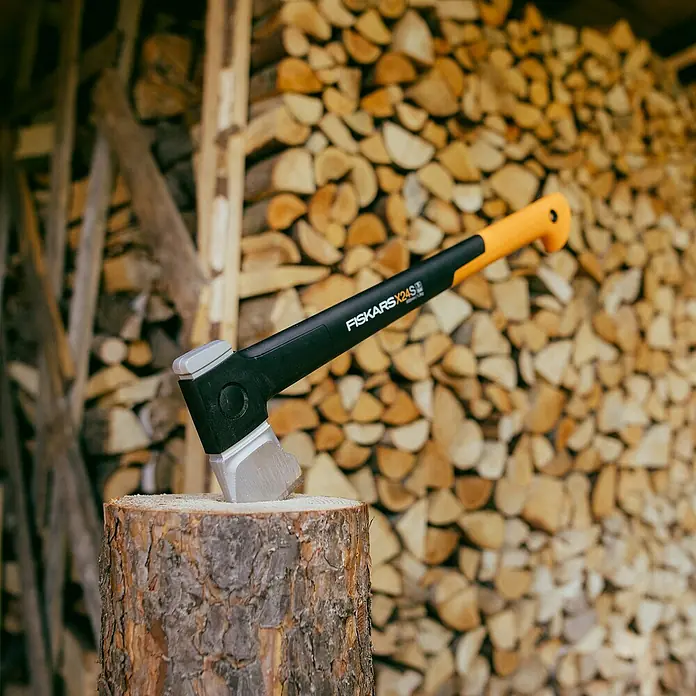 Fiskars X-series Spaltaxt X24 S - Länge: 60 cm - Nahaufnahme einer Fiskars X24 Spaltaxt, die in einem Holzklotz steckt, mit einem Stapel Brennholz im Hintergrund.