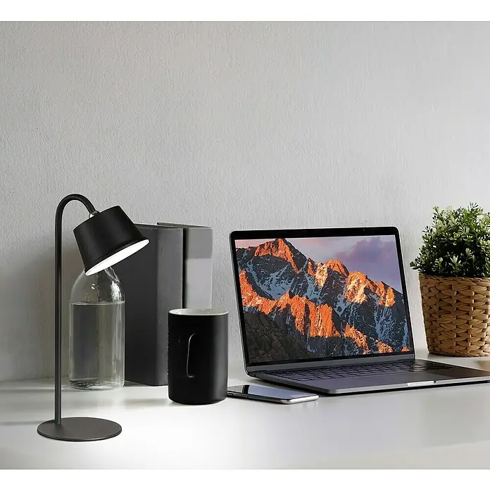 FHL easy Akku-Tischleuchte VOET - 3 W, L x B x H: 17 x 12 x 34 cm, Sandschwarz, Warmweiß - Der schwarze Schreibtischlampe spendet Licht neben einem Laptop, einer Tasse, Büchern und einer Pflanze auf einem weißen Schreibtisch.
