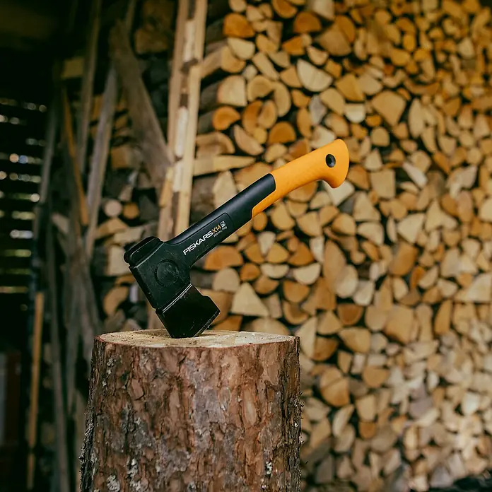 Fiskars X-series Universalaxt X14 - Länge: 35,5 cm - Axt Fiskars X14 auf einem Holzklotz, mit einem Hintergrund aus gestapeltem Brennholz. Ideal für Gartenarbeiten und Holzarbeiten.