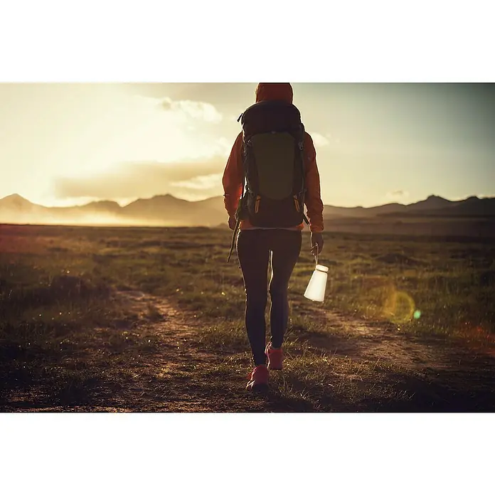 FHL easy Akku-Tischleuchte BRUNO - 0,5 W, L x B x H: 12 x 12 x 20 cm, Weiß, RGB - Frau wandert mit Rucksack und einer tragbaren Laterne in der Hand durch eine Landschaft im goldenen Licht der Abenddämmerung.