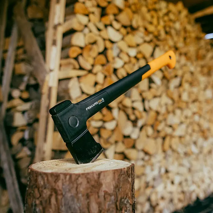 Fiskars X-series Universalaxt X24 M - Länge: 60 cm - Nahaufnahme einer Fiskars X24 Spaltaxt, die in einen Holzscheit gesteckt ist, mit einem Stapel Brennholz im Hintergrund.
