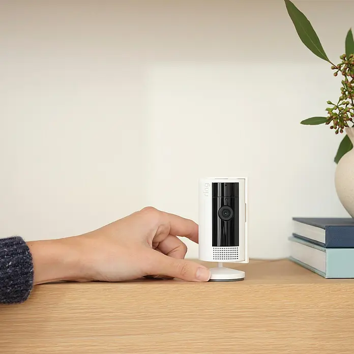 Ring Innenkamera Indoor Cam 2. Gen. - 1.080 Pixel (Full HD), Weiß, 1 Stk. - Nahaufnahme einer weißen Ring-Überwachungskamera auf einem Holztisch, die mit der Hand bedient wird, mit minimalistischem Design.