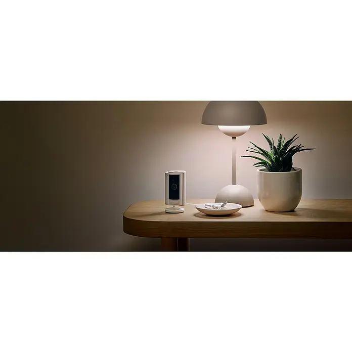 Ring Innenkamera Indoor Cam 2. Gen. - 1.080 Pixel (Full HD), Weiß, 1 Stk. - Ein Ring Stick Up Cam batteriebetrieben, eine Lampe und eine Pflanze stehen auf einem Tisch. Der Fokus liegt auf der Ring Überwachungskamera.