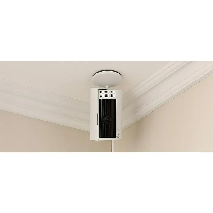 Ring Innenkamera Indoor Cam 2. Gen. - 1.080 Pixel (Full HD), Weiß, 1 Stk. - Nahaufnahme einer weißen Ring-Überwachungskamera, die an einer weißen Decke mit Zierleisten montiert ist und Sicherheit bietet.