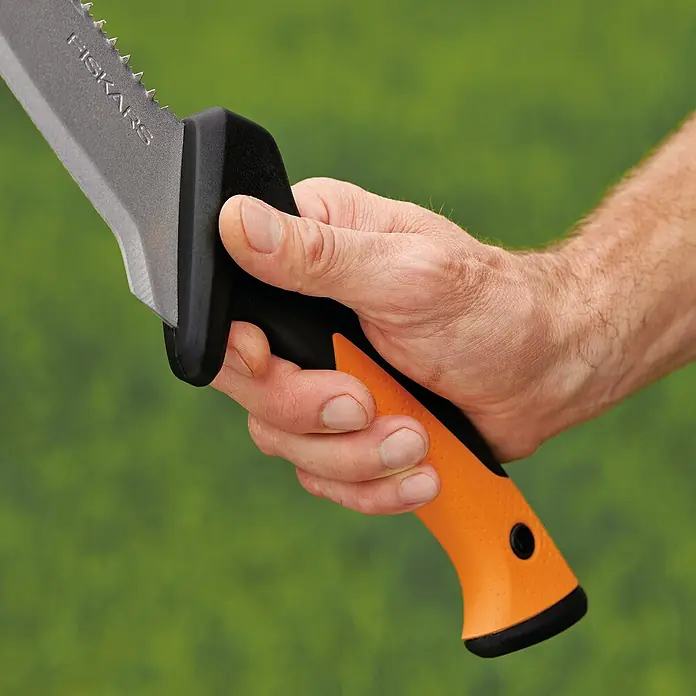 Fiskars Solid Machete mit Sägekante - Länge: 45 cm - Nahaufnahme einer Hand, die eine Fiskars Machete mit orangefarbenem Griff vor einem grünen Hintergrund hält.