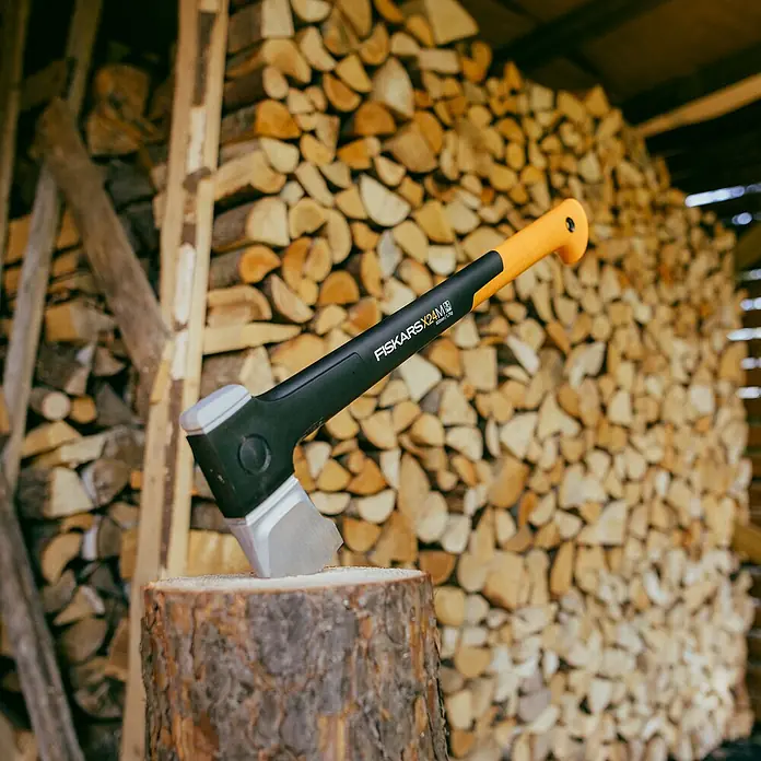 Fiskars X-series Spaltaxt X24 M - Länge: 60 cm - Nahaufnahme einer Axt der Marke Fiskars, Modell X24M, die in einem Holzscheit steckt, mit einem Stapel Holz im Hintergrund.