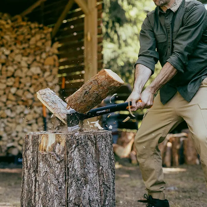 Fiskars X-series Spaltaxt X28 - Länge: 71 cm - Ein Mann spaltet Holz mit einer Fiskars X25 Axt auf einem Hackklotz, Holzspäne fliegen herum. Im Hintergrund ist ein Holzschuppen zu sehen.