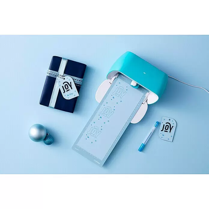 Cricut Joy Schneideplotter Starter Bundle - Türkis/Weiß, Bluetooth, Schnittbreite: 10,4 cm, Schnittlänge: 1,2 m - Eine aquafarbene Cricut Joy schneidet hellblaue Matten mit 'JOY'-Geschenkanhängern und Schneeflocken. Daneben ein verpacktes Geschenk und Ornamente.