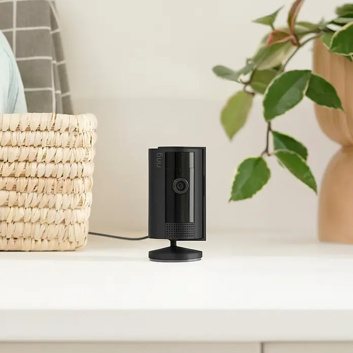 Ring Innenkamera Indoor Cam 2. Gen. - 1.080 Pixel (Full HD), Schwarz, 2 Stk. - Nahaufnahme einer schwarzen Ring Sicherheitskamera, die auf einer weißen Oberfläche steht, mit einem Korb und einer Pflanze im Hintergrund.