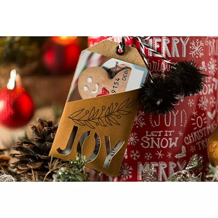 Cricut Joy Schneideplotter Smarte Schneide- und Zeichenmaschine - Türkis/Weiß, Bluetooth, Schnittbreite: 10,4 cm, Schnittlänge: 1,2 m - Geschenkkartenhalter aus Kraftpapier mit 'JOY'-Ausschnitt und Lebkuchenmann-Geschenkkarte, umgeben von Weihnachtsdeko.