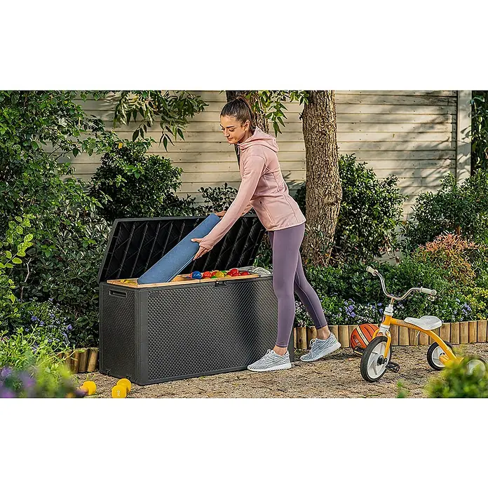 Keter Arcón para cojines de exterior Emily - 117,8 x 44,6 x 58 cm, Plástico, Gris - Una mujer guarda una esterilla de yoga azul en una caja de jardín negra, al lado de la cual hay un triciclo amarillo.