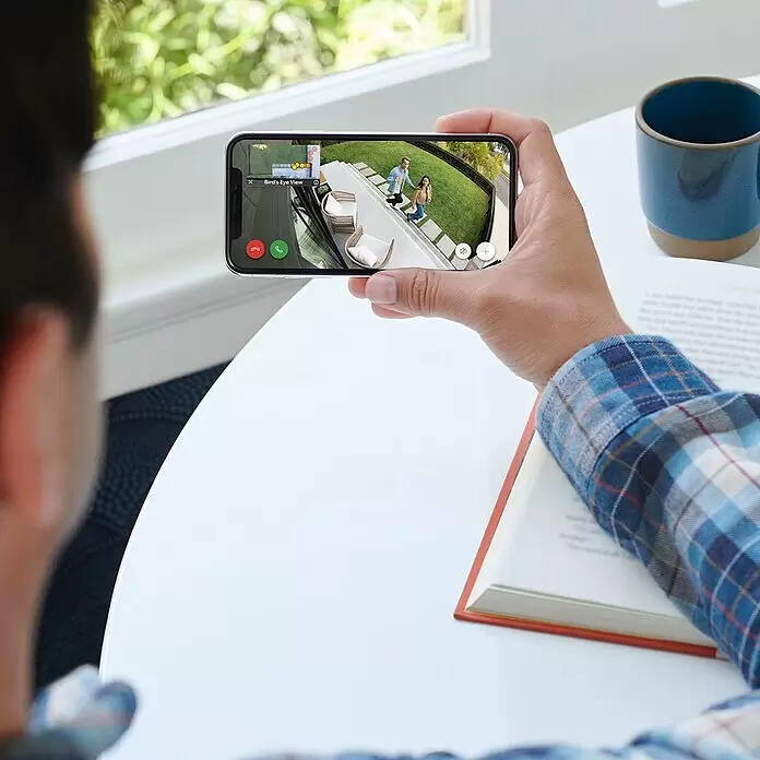 Ring Cámara de vigilancia Ring Spotlight Cam Pro Wired - Blanco, Ángulo de detección: 140 °, Funcionamiento en red - Una persona sostiene un smartphone negro horizontalmente, viendo una transmisión en vivo de cámara de seguridad con dos personas en un césped con muebles de patio.