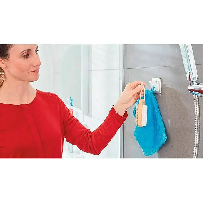 Tesa Powerstrips Doppelhaken Large - 1 Stk., Metall - Frau hängt Handtuch und Bürste an einem weißen Wandhaken im Badezimmer auf. Praktische Badaccessoires für den täglichen Gebrauch.