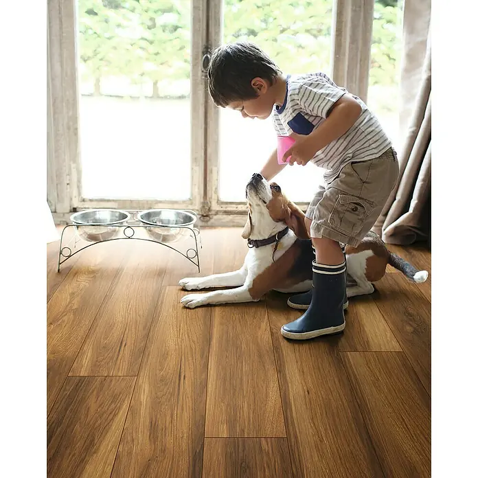 Krono Original Laminat Atlantic 8 - Silverdale Oak, Landhausdiele, 1.285 x 192 x 8 mm - Ein Junge in Stiefeln gibt einem Hund, der auf einem Holzboden liegt, mit einem rosa Becher Wasser. Eine Doppel-Futterstation aus Edelstahl steht daneben.