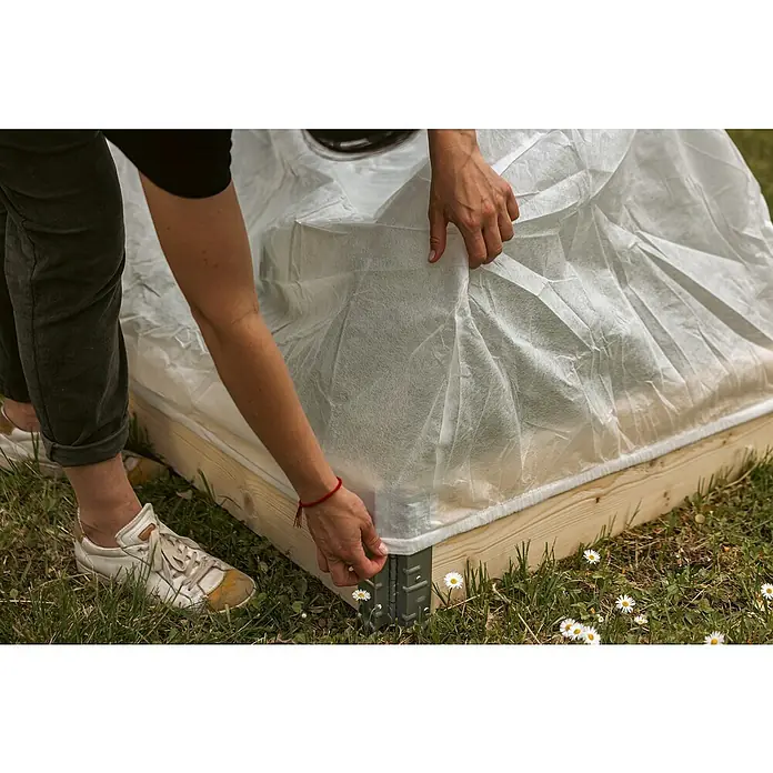 Upyard Abdeckvlies - 120 x 80 cm, Passend für: Bogen Hochbeetumrandung - Eine Person befestigt eine weiße Gartenabdeckung an einem Hochbeet aus Holz mit Metallwinkeln im Rasen.
