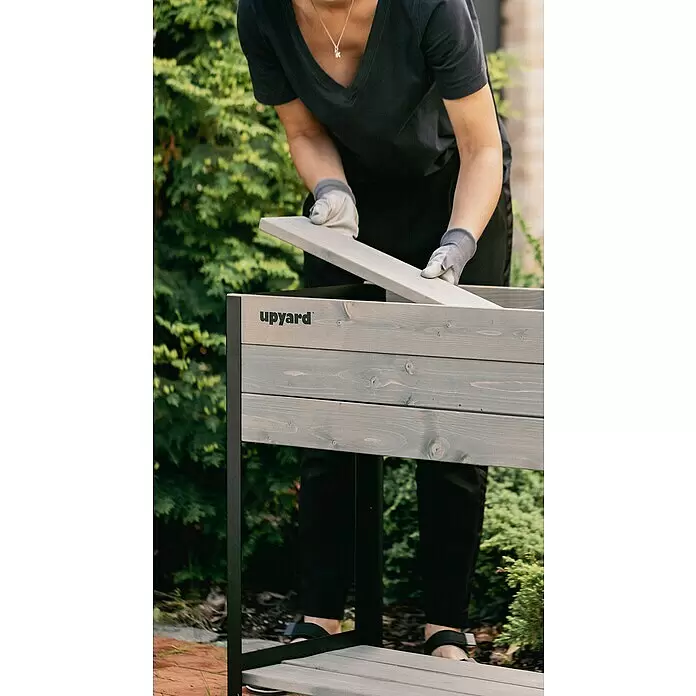 Upyard Huerto Modernbox alto - 118 x 36 x 72 cm, Madera, Gris/Negro - Una persona ensambla una jardinera elevada de madera gris con estructura de metal negro, colocando una tabla.