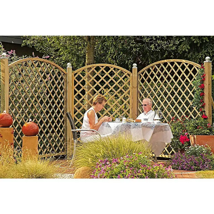 T & J Zaunelement BogenrankenB x H: 180 x 180 cm, Holz, Maschenbreite: 10 cm - Gartenszene mit hölzernem Sichtschutzzaun mit Rautenmuster, der eine private Sitzecke mit Tisch und Paar umrahmt, umgeben von üppigem Grün.