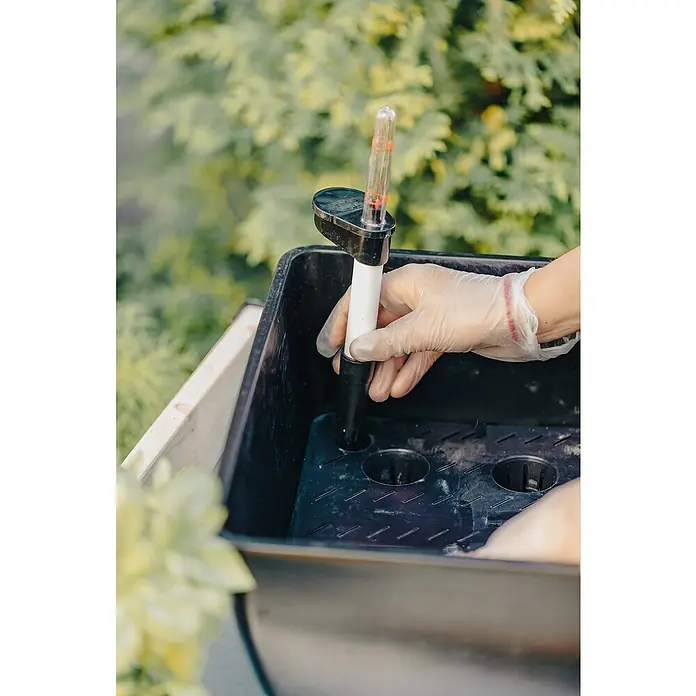Upyard BewässerungssystemPassend für: Holzaufsatzrahmen 80 x 60 cm - Eine Hand mit Handschuh setzt einen Füllstandsanzeiger in ein schwarzes Pflanzgefäß, um den Wasserstand zu prüfen. Gartengerät im Einsatz.