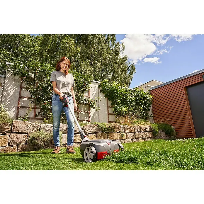 AL-KO Bosch Home & Garden Compatible Akku-Rasenmäher 22.4 LI Comfort - 18 V, 1 Akku, 2,5 Ah - Eine Frau bedient einen Einhell Akku-Rasenmäher im Garten, umgeben von üppigem Grün und einer Steinmauer, unter blauem Himmel.