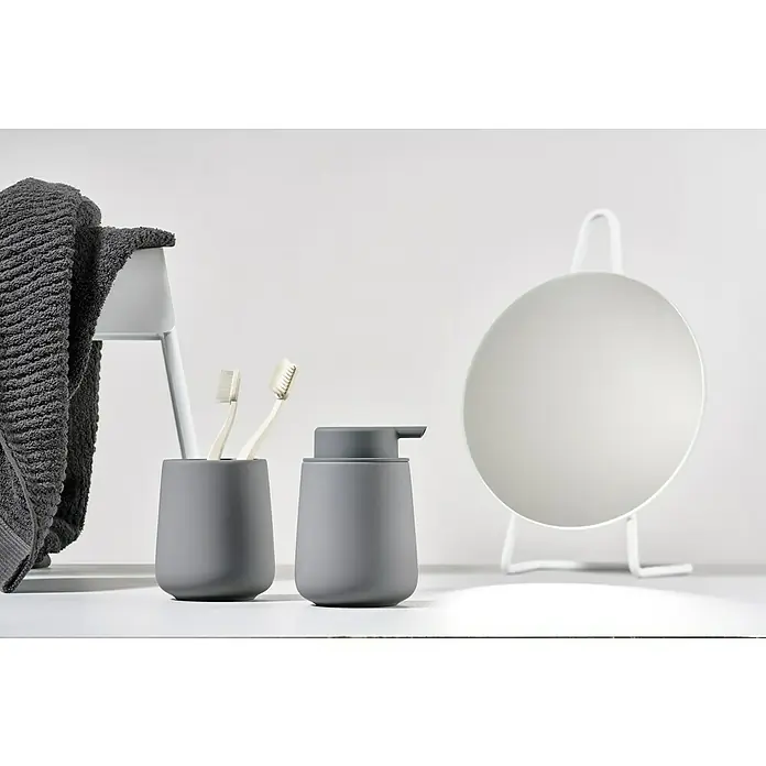 Zone Denmark Nova One Zahnputzbecher - Keramik, Gull Grey - Badezimmer-Arrangement mit grauem Handtuch, Zahnbürsten, Seifenspender und einem runden Spiegel mit weißem Rahmen auf einem weißen Tisch.