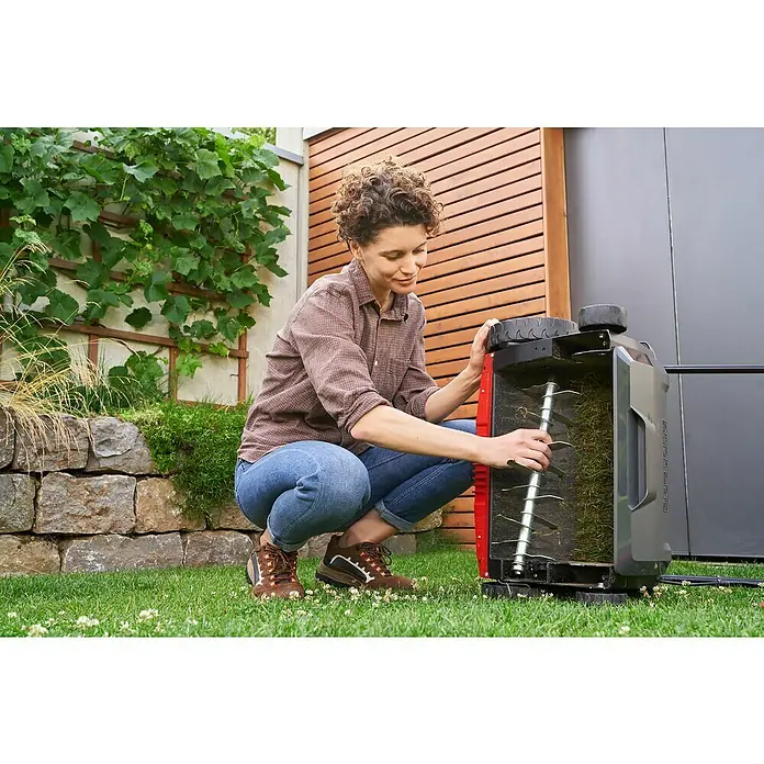 AL-KO Easy Elektro-Vertikutierer-Lüfter 38.4 E Combi Care - 1.400 W, Arbeitsbreite: 38 cm - Eine Frau hockt neben einem Rasenlüfter und inspiziert die Walze. Sie trägt Jeans, ein Hemd und braune Schuhe.
