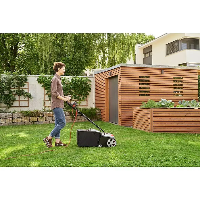AL-KO Easy Elektro-Vertikutierer-Lüfter 38.4 E Combi Care - 1.400 W, Arbeitsbreite: 38 cm - Eine Frau benutzt einen Rasenmäher auf einem Rasen vor einem Holzgebäude.