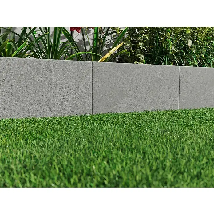 Diephaus L-Stein - 30 cm, 50 cm, Stärke: 7 cm, Grau - Detailaufnahme von grauen Rasenkantensteinen, die eine grüne Rasenfläche von einem Gartenbeet mit üppiger Bepflanzung abgrenzen.