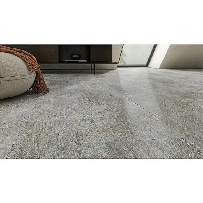 Feinsteinzeugfliese Saloon Silver - 30 x 60 cm, Grau/Silber, Matt - Nahaufnahme eines grauen Holzfußbodens mit einem Pouf und einer hellbraunen Decke im Hintergrund, der die Textur und das Design hervorhebt.