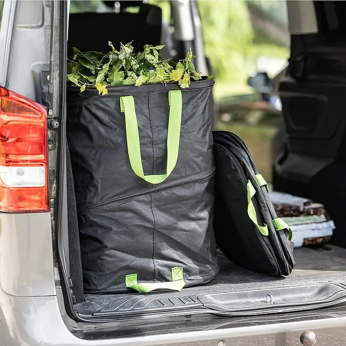 Windhager Pop-Up Gartenabfallsack Square - Fassungsvermögen: 180 l, B x H: 52 x 68 cm, Schwarz - Ein schwarzer Gartensack mit grünen Griffen, voll mit Blättern, steht im Kofferraum eines Autos. Daneben liegt ein zusammengefalteter Sack.