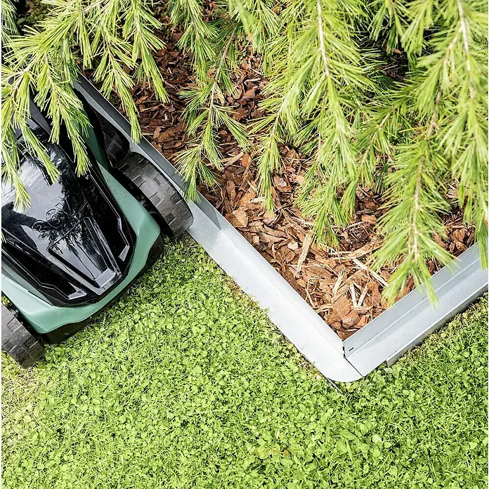 Windhager Rasenkante Easycut - L x B x H: 80 x 0,4 x 11 cm, Stahl, Verzinkt - Ein Rasenmähroboter mäht den Rasen neben einem erhöhten Gartenbeet mit grünen Pflanzen und Mulch. Die Rasenfläche ist sattgrün.