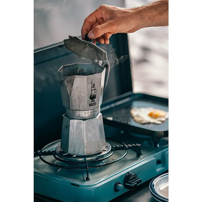 Cadac Grillständer für Kaffeekannen - Passend für: Cadac 2 Cook / Safari Chef 30 / Citi Chef 40 / Carri Chef 40; Topfträger mit 3 oder 4 Beinen - Bialetti Espressokocher auf Campingkocher mit Hand am Deckel, dampfend. Perfekt für Kaffee unterwegs, mit Ei in der Pfanne daneben.