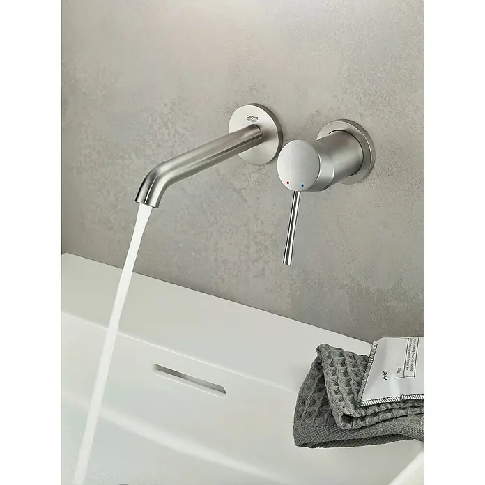 Grohe Essence Waschtischarmatur L - Wandmontage, Supersteel, Matt - Nahaufnahme einer modernen Grohe Waschtischarmatur aus Edelstahl mit Wasserfluss, montiert über einem weißen Waschbecken.