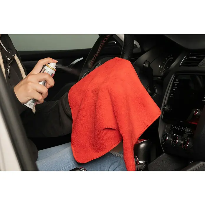Nigrin Performance Display-Reiniger - 75 ml - Innenraumreinigung: Eine Person sprüht Reinigungsmittel auf ein rotes Tuch, um das Armaturenbrett eines Autos zu reinigen.