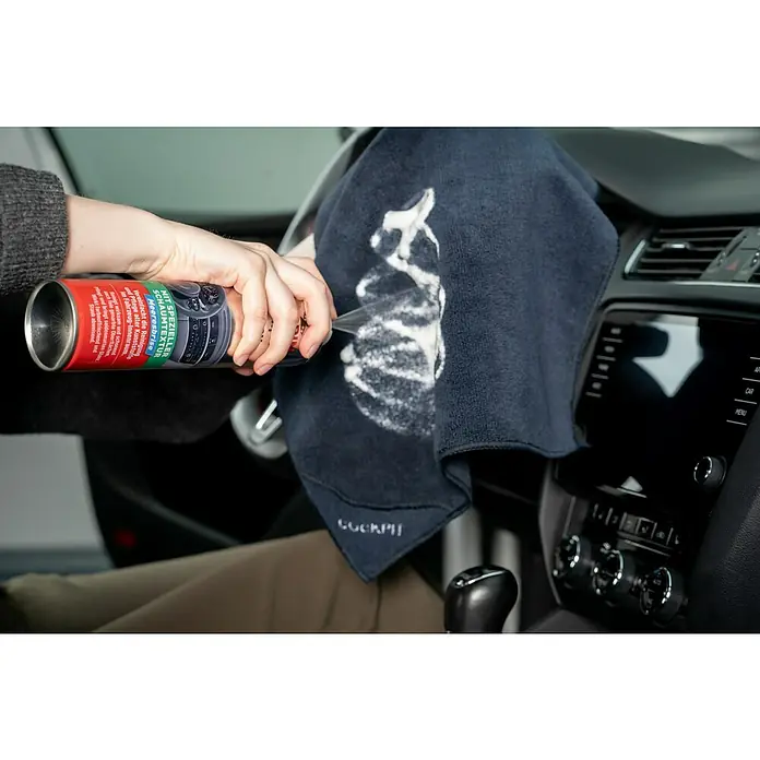 Nigrin Cockpit-Spray - 400 ml, Vanille - Detailaufnahme einer Person, die Cockpitreiniger-Schaumspray auf ein Tuch sprüht, um das Armaturenbrett eines Autos zu reinigen.