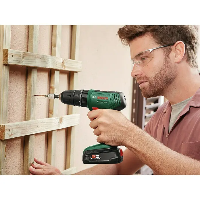 Bosch Power for All 18V Akku-Schlagbohrschrauber EasyImpact 18V-38 - 18 V, 1 Akku, 2 Ah, 38 Nm - Mann benutzt einen grünen Bosch EasyImpact 18V-38 Akku-Bohrschrauber, um Holzleisten an einer Wand zu befestigen, und trägt dabei eine Schutzbrille.