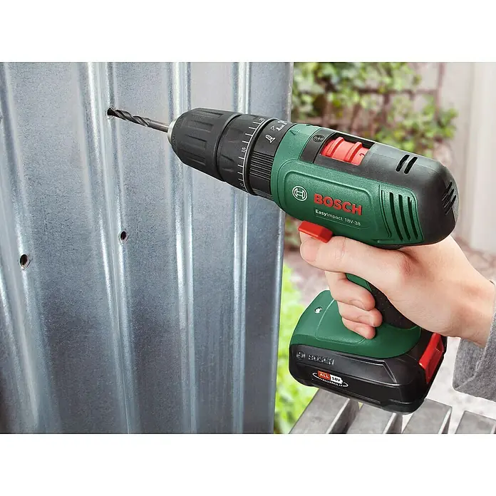 Bosch Power for All 18V Akku-Schlagbohrschrauber EasyImpact 18V-38 - 18 V, 1 Akku, 2 Ah, 38 Nm - Akkuschrauber von Bosch beim Bohren eines Lochs in eine Metallplatte. Das Werkzeug ist grün und schwarz und hat einen roten Knopf.